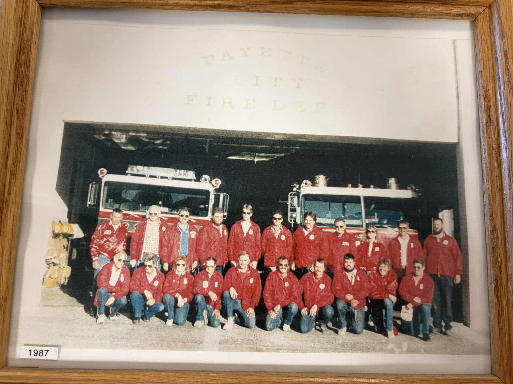 1987 Payette City Fire Department crew in red jackets in front of two engines in the station bay