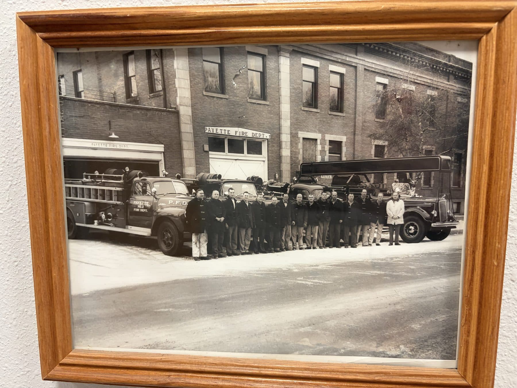 Black and white photo of Payette firefighters lined up in front of the old downtown brick fire station with two engines and a ladder truck