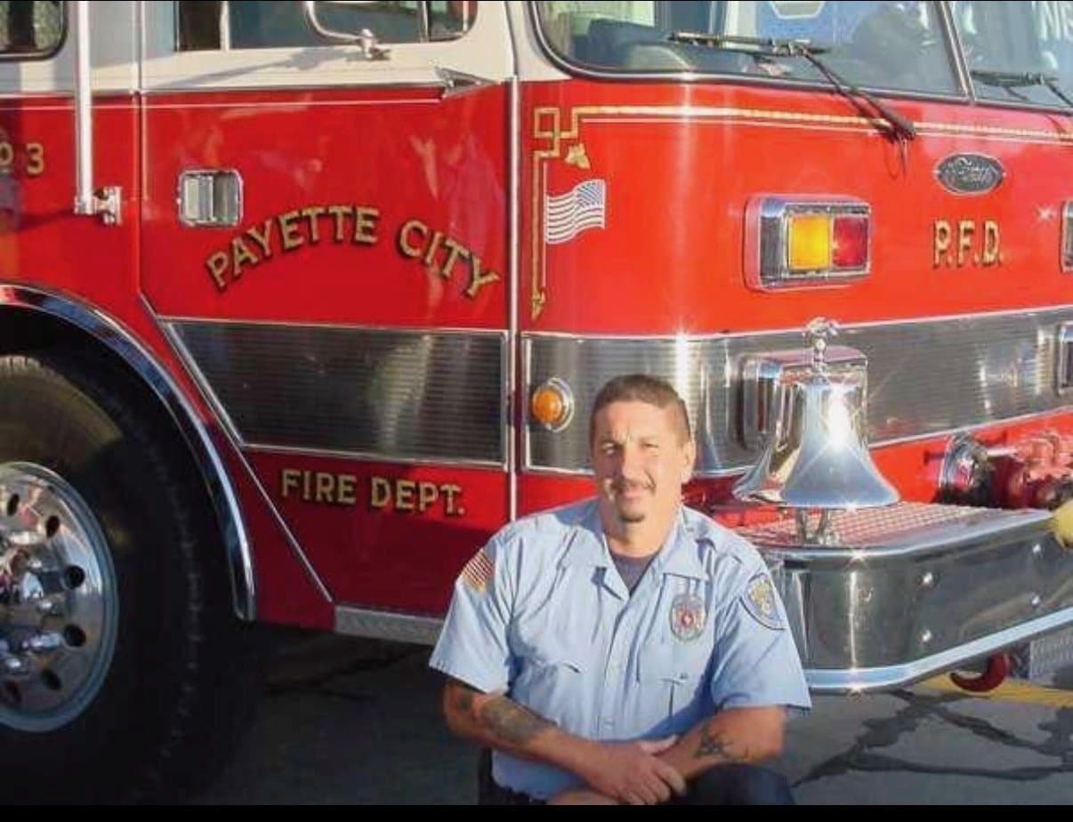 A young Steve Castenada in uniform, kneeling in front of a Payette City Fire Dept. engine early in his career